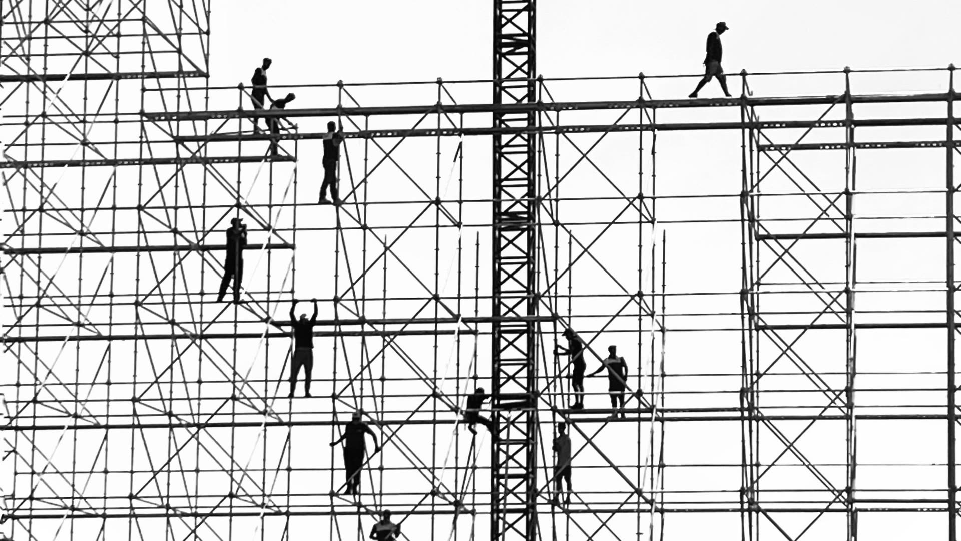 Gerüstbauer als Silhouetten gegen den hellen Himmel auf einem mehrgeschossigen Gerüst, einige laufen über die Belaglagen, andere klettern zwischen den Rahmen