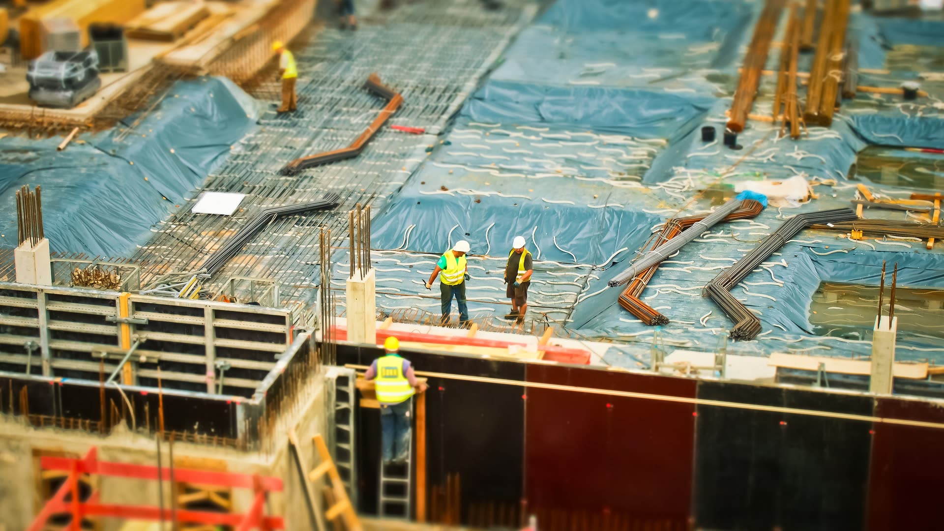 Blick von oben auf eine Großbaustelle: Arbeiter in Warnwesten auf einer mit Folie abgedeckten Bewehrungslage, daneben gebündelter Bewehrungsstahl