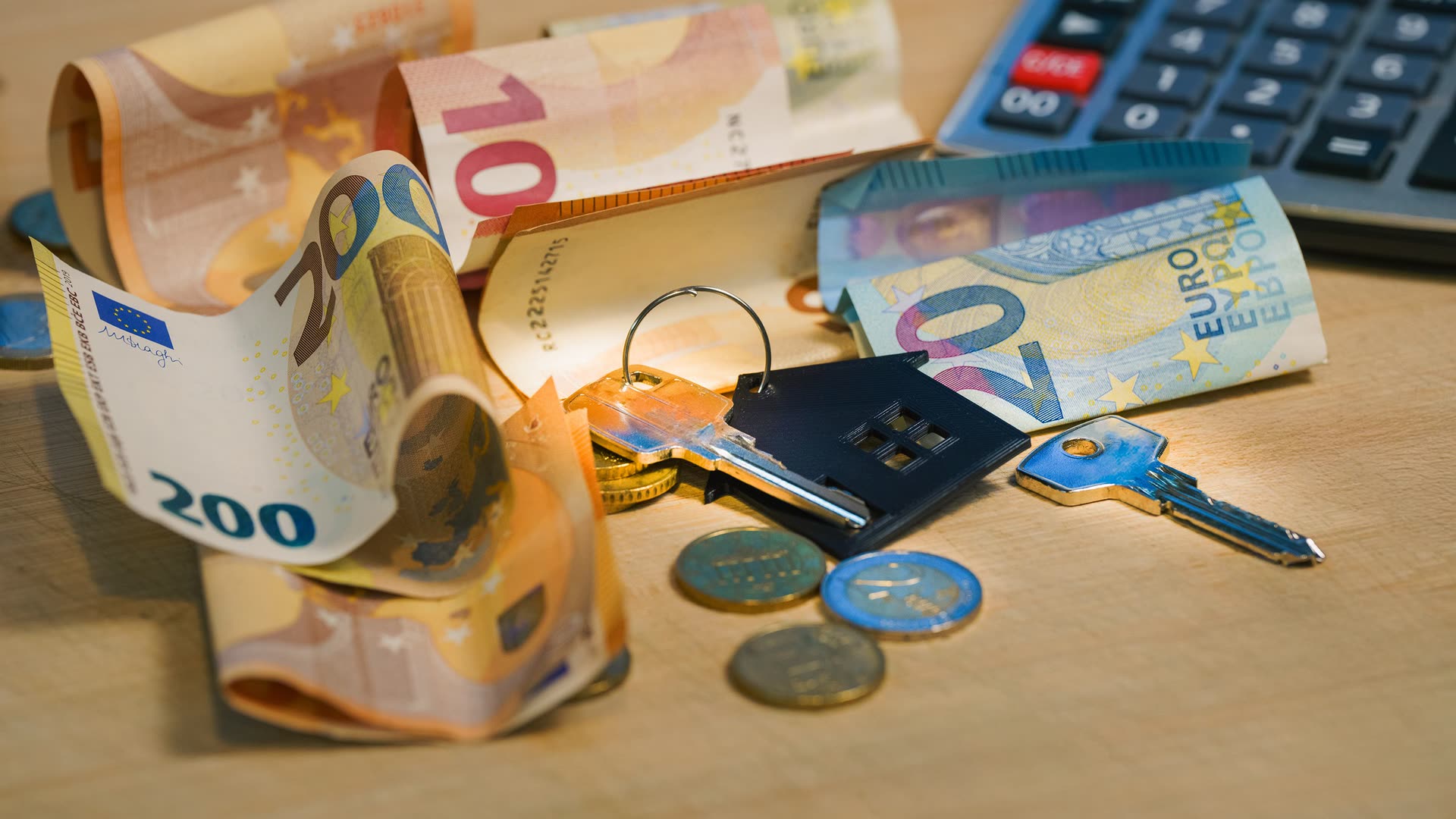 Euro-Geldscheine, Münzen, ein Hausschlüssel mit Haus-Anhänger und ein Taschenrechner liegen auf einem hellen Holztisch